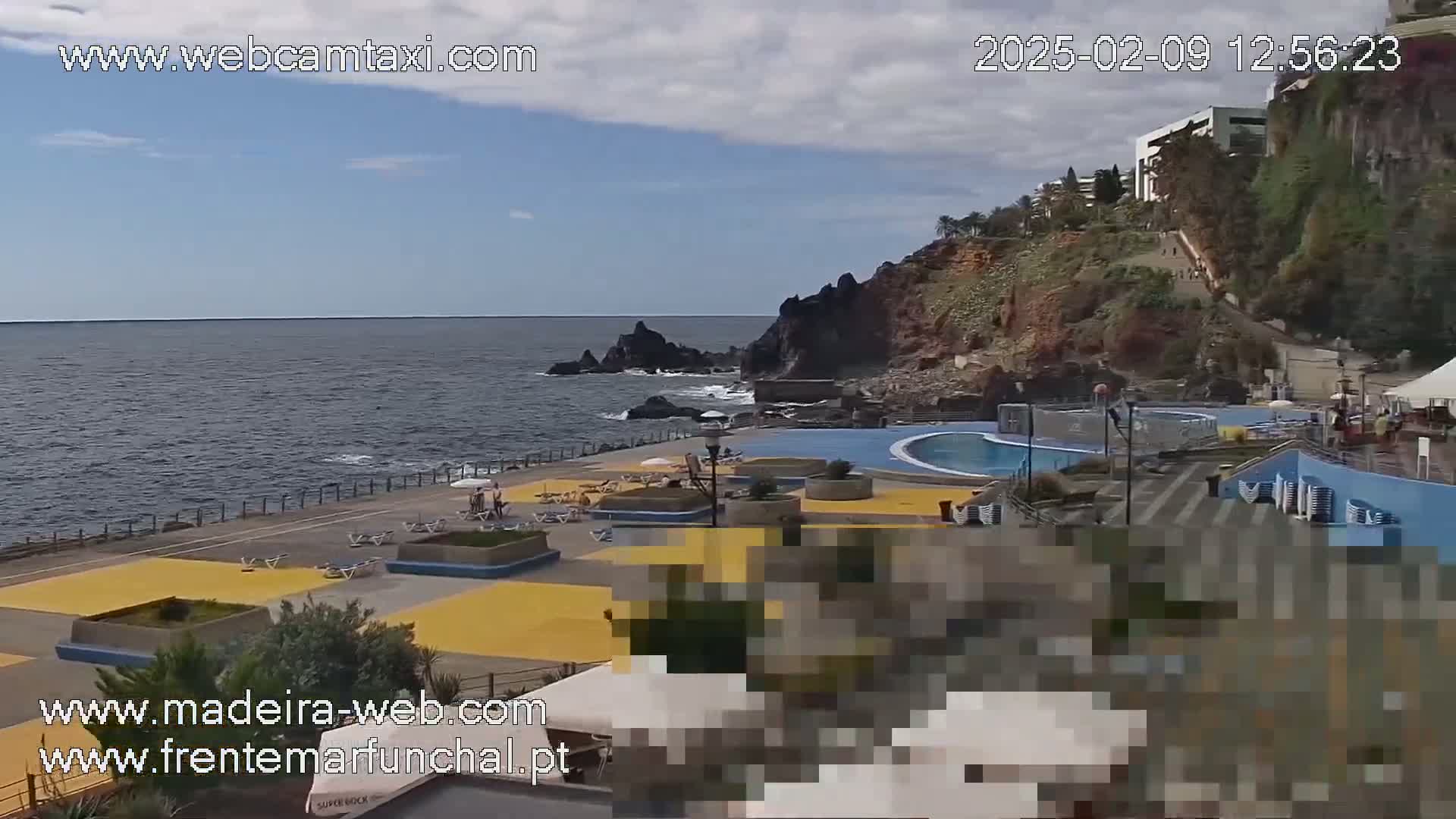 A mostly sunny outdoor scene shows a seaside resort with pools and a walkway next to dark volcanic rock cliffs and a calm ocean.