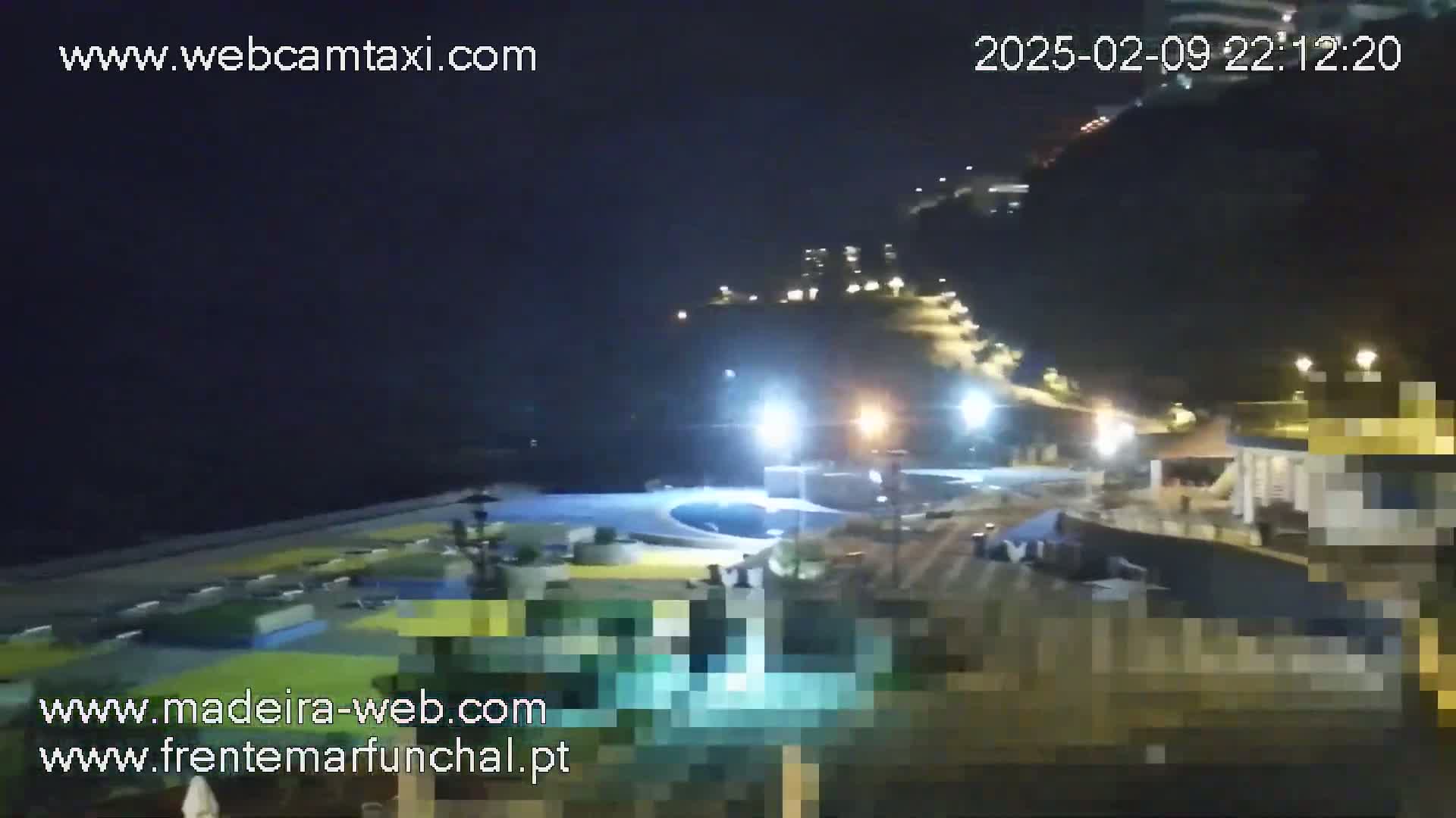 A nighttime view of a coastal area with buildings, lights, and a swimming pool, under a clear, dark sky.