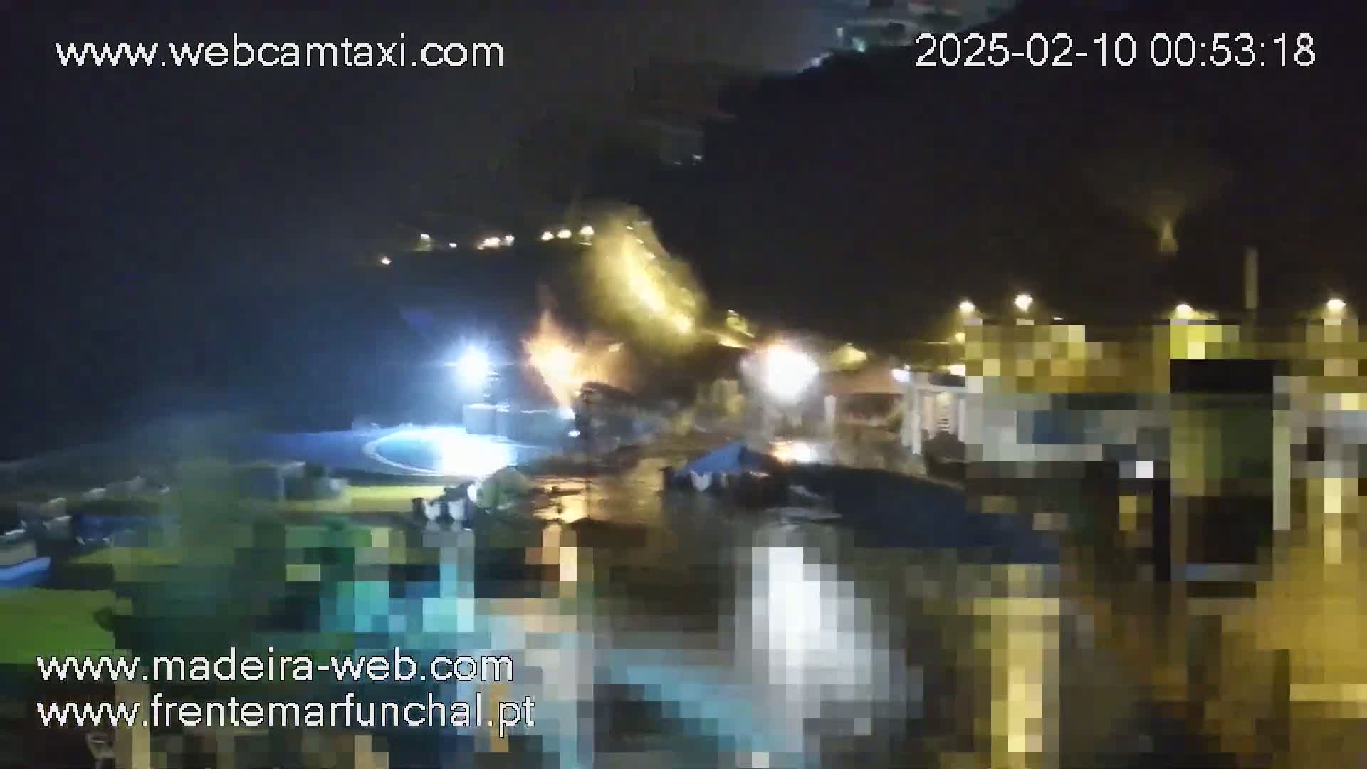 A nighttime, illuminated view of a waterfront area with buildings, a pool, and some boats, partially obscured by rain or mist.