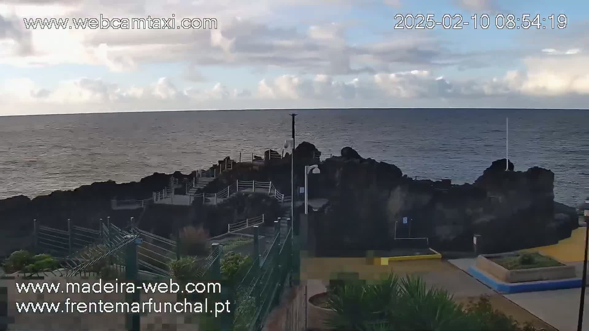 A rocky coastal area with stairs leading down to the ocean is viewed under a partly cloudy sky.