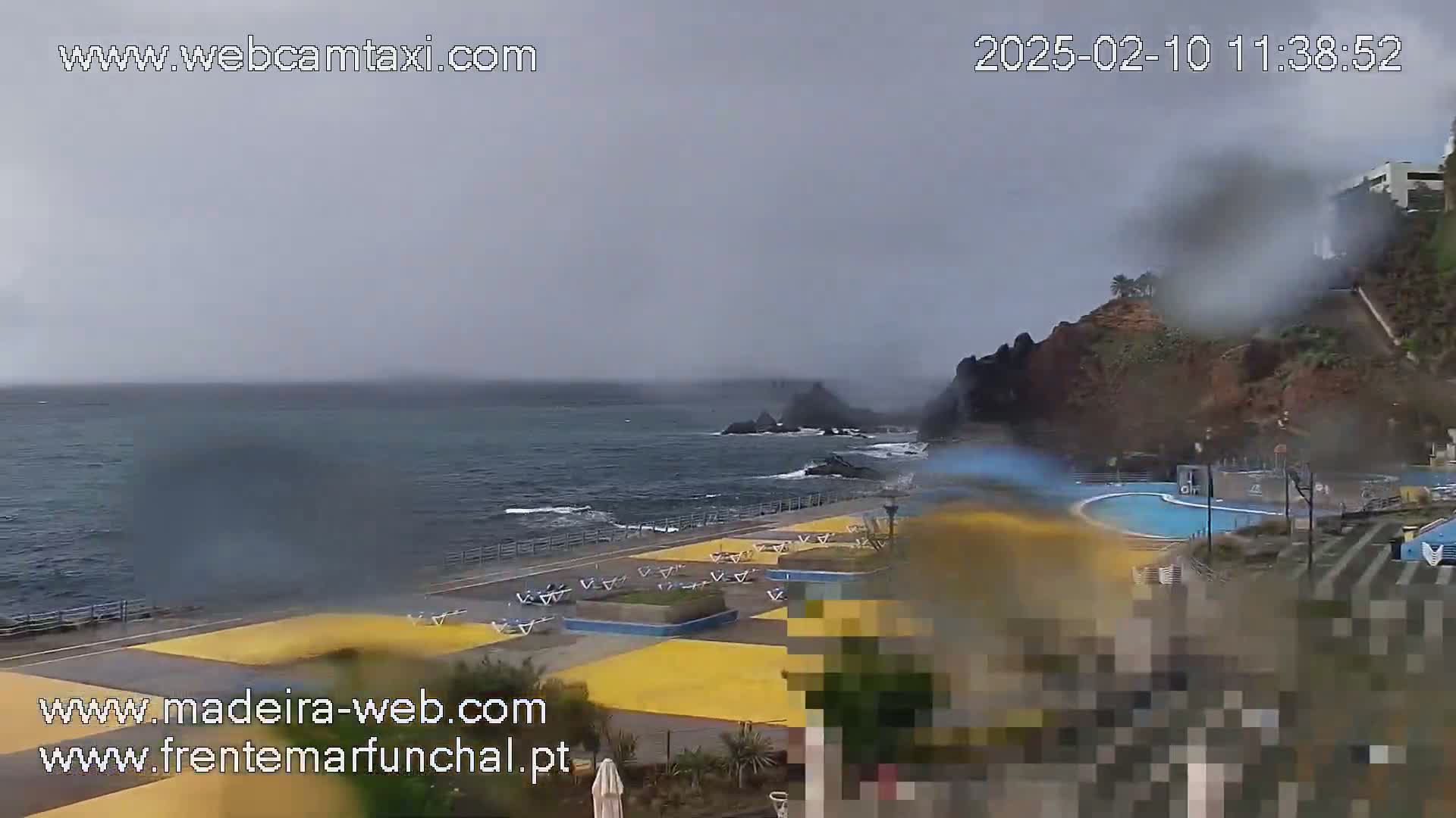 A rain-swept seaside view shows a swimming pool and empty lounge chairs along a paved walkway near dark, rocky cliffs and a rough ocean.