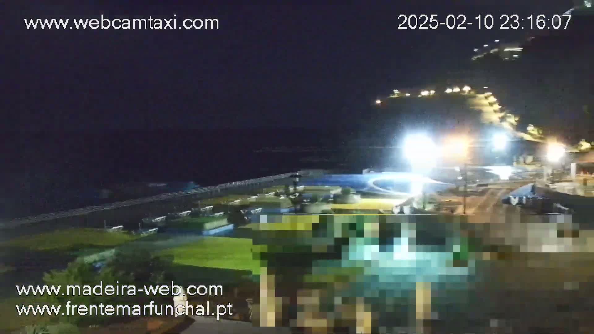 A nighttime view of a coastal area with brightly lit buildings and a dark ocean under a clear sky.