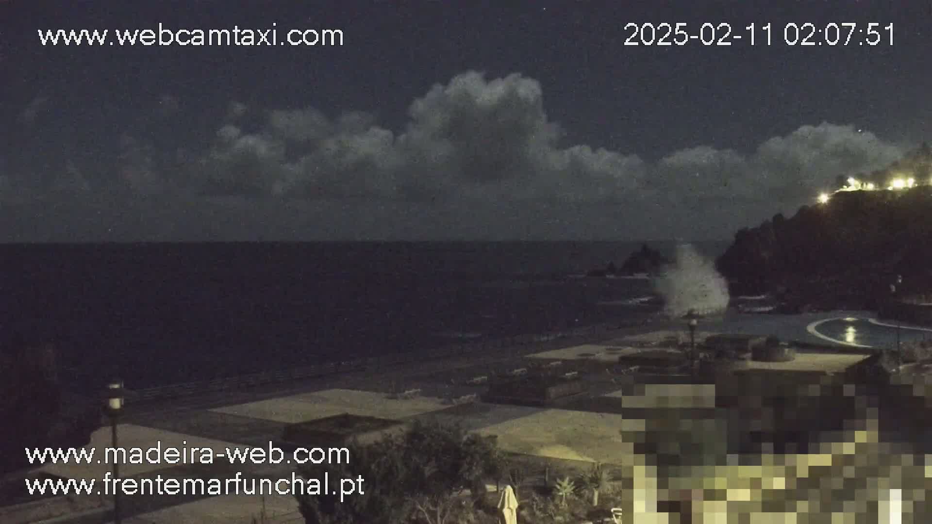 A nighttime coastal scene shows a dark ocean with waves crashing against rocks near a shoreline with some buildings and vegetation under a partly cloudy sky.