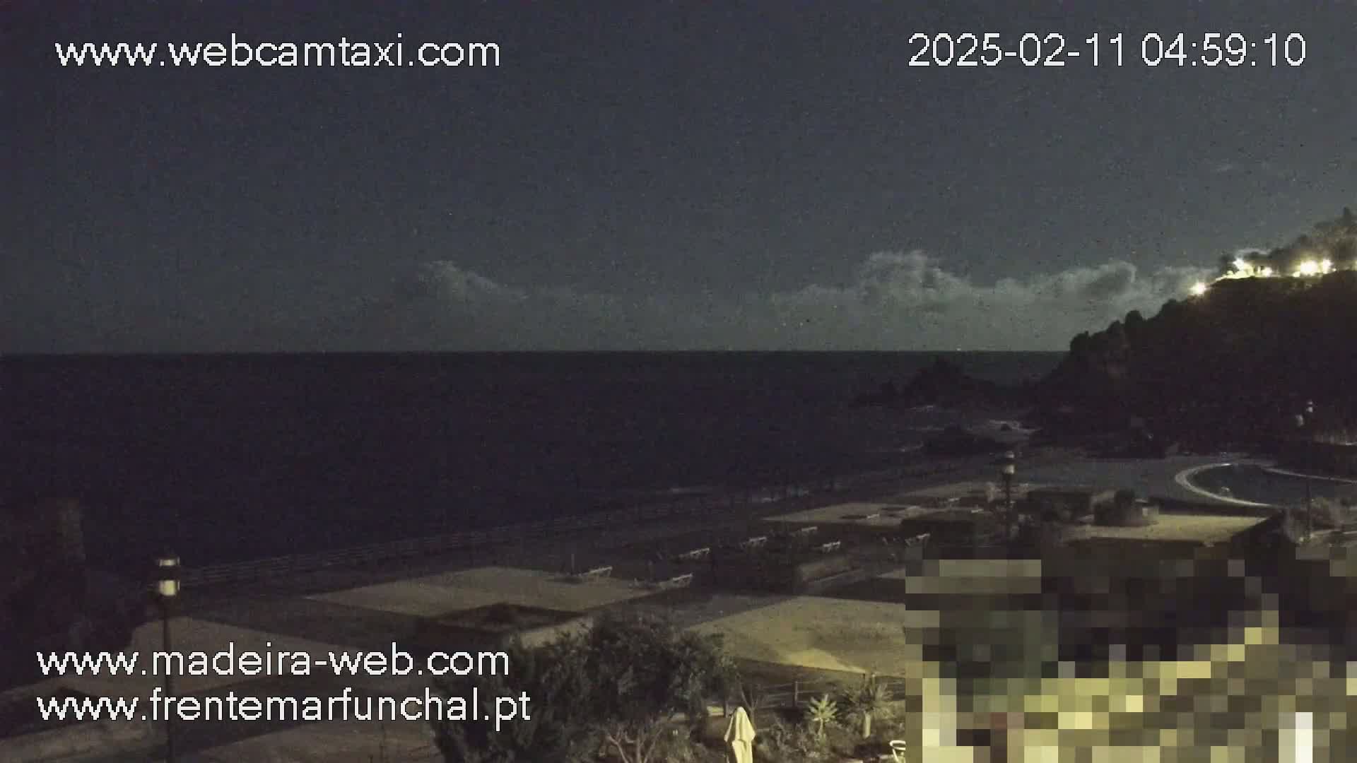 A nighttime view of a dark ocean under a partly cloudy sky shows a shoreline with some buildings and landscaping.
