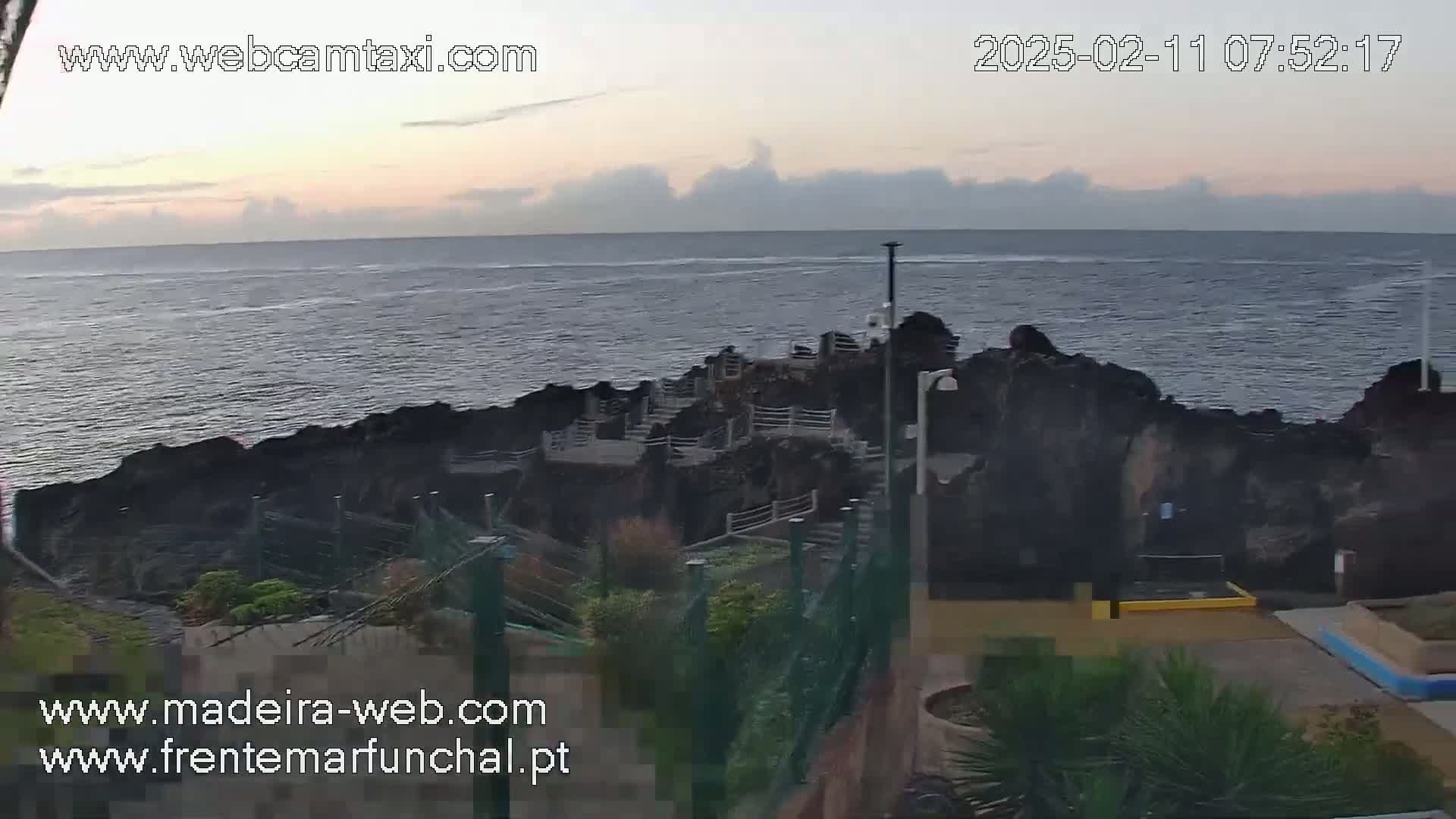 A rocky coastline with a walkway overlooks a calm ocean under a mostly clear, dusky sky.