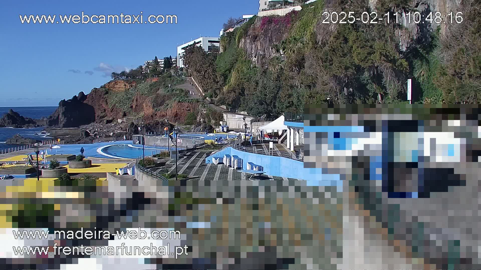An outdoor oceanside swimming pool complex sits on a rocky cliff overlooking the ocean on a sunny day.