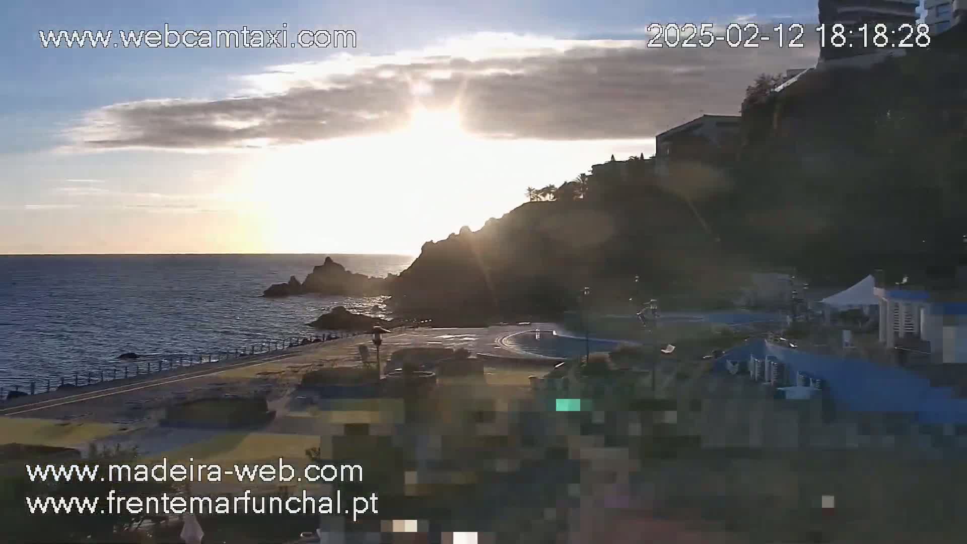 A seaside scene at sunset, featuring a rocky cliff with buildings on top overlooking a calm ocean and a paved area with structures below.