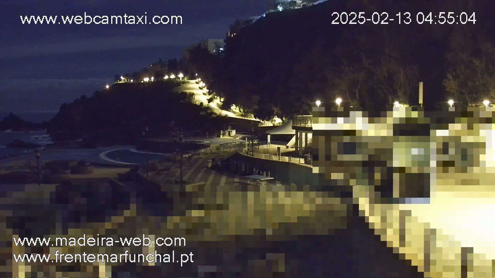 A nighttime view of a coastal area with a paved walkway illuminated by lights winding up a hillside toward buildings, overlooking a dark body of water.