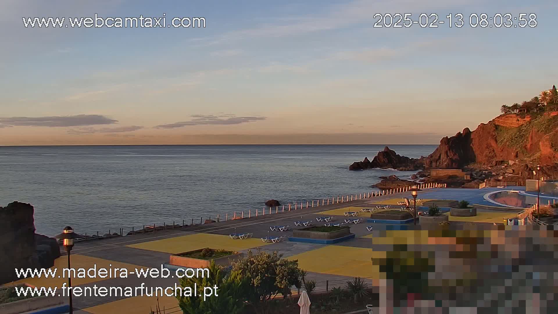A calm ocean at sunrise overlooks a paved seaside area with lounge chairs and a pool, backed by reddish-brown cliffs.