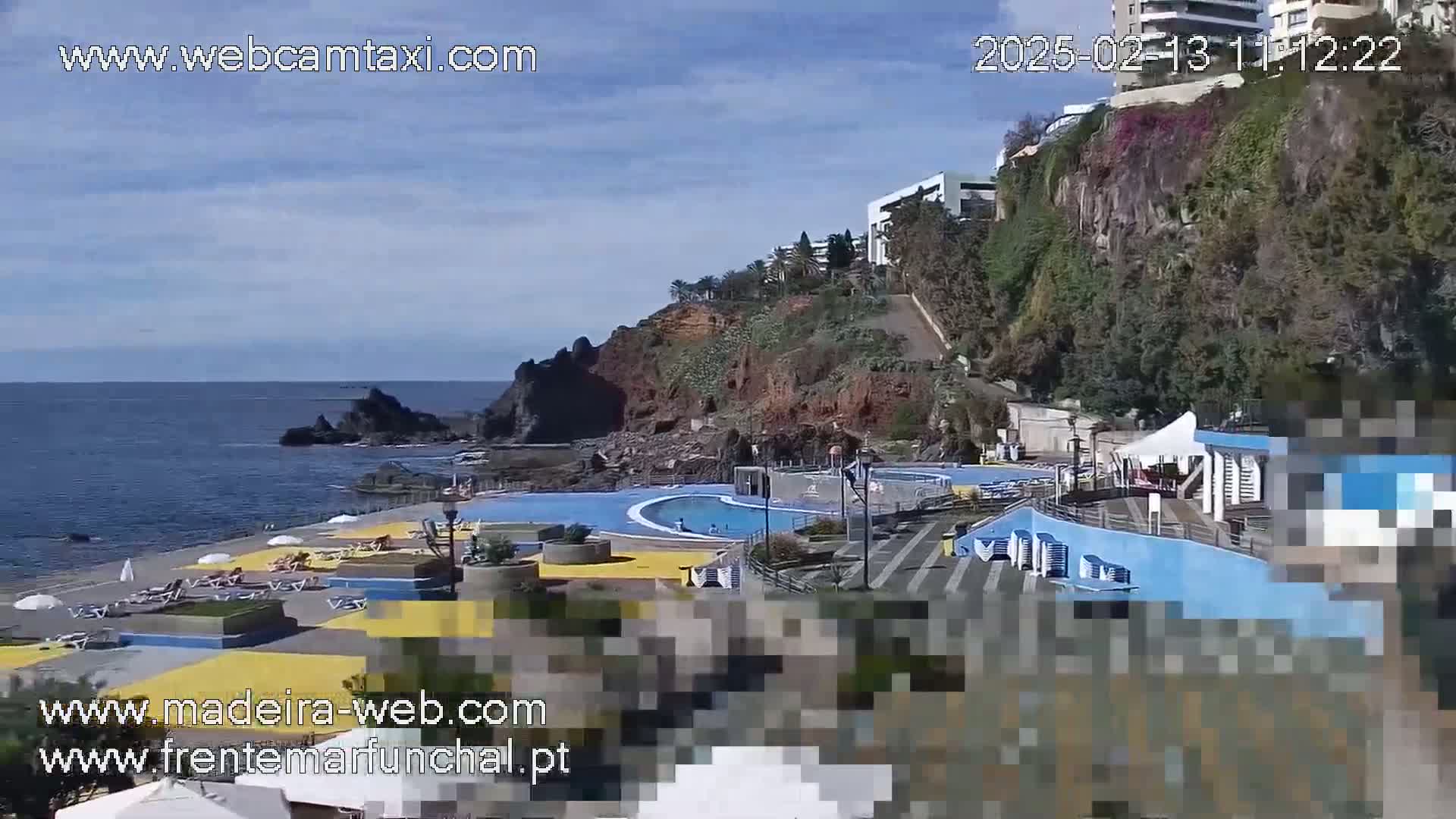 An outdoor scene shows a coastal area with a large pool, beach, and buildings on a cliff overlooking the ocean under a mostly clear sky.