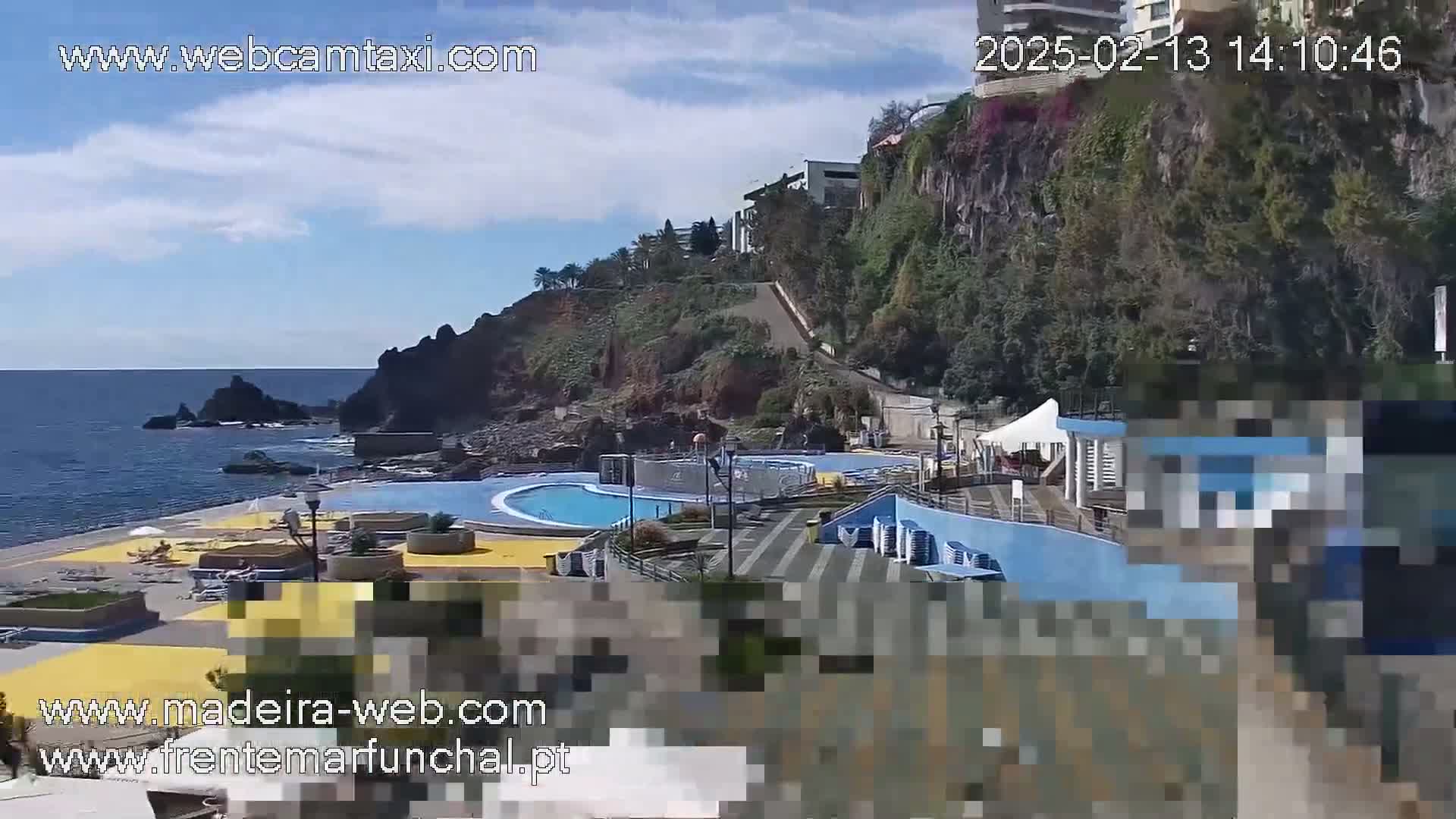 An outdoor scene shows a swimming pool complex situated on a rocky coastline overlooking a calm ocean under a partly cloudy sky.