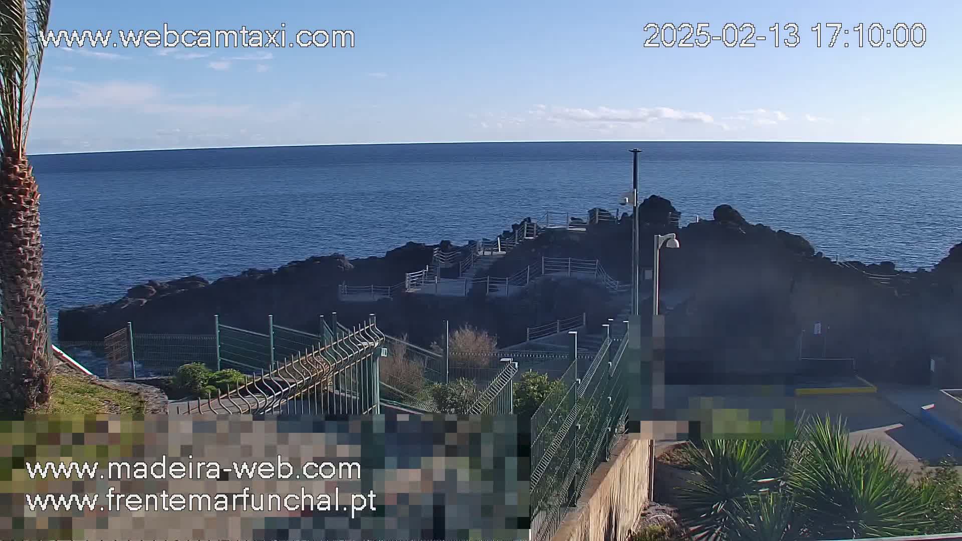 A rocky coastal area with a walkway and fences overlooks a calm ocean under a mostly clear sky.