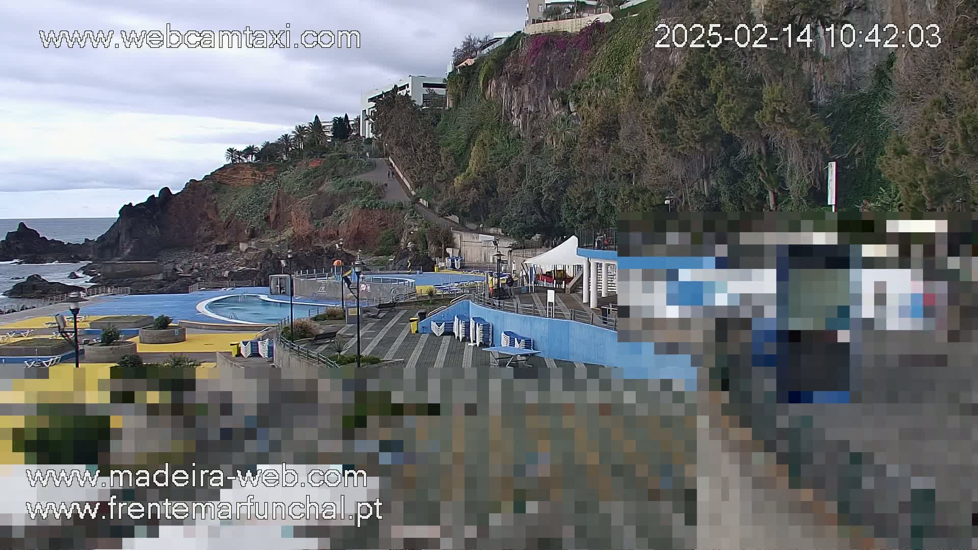 An outdoor swimming pool complex is situated on a rocky coastline under a cloudy sky.