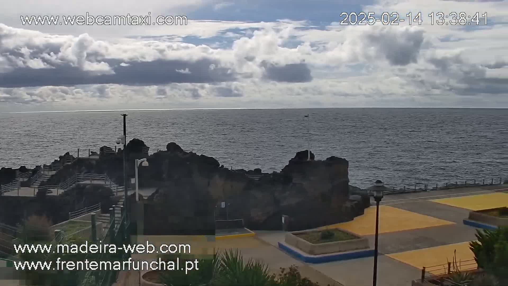 A partly cloudy day reveals a dark-colored ocean meeting a rocky shoreline with a paved walkway and planted areas.