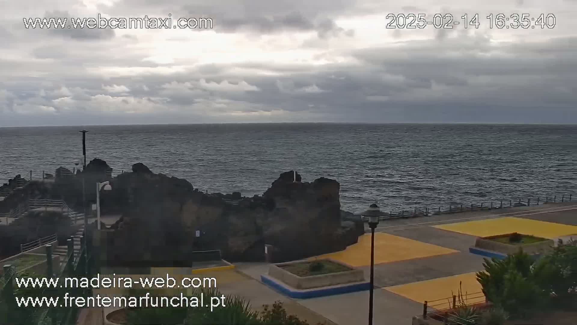A rocky coastline with a paved area featuring yellow sections is visible under a cloudy sky, overlooking a dark-blue ocean.