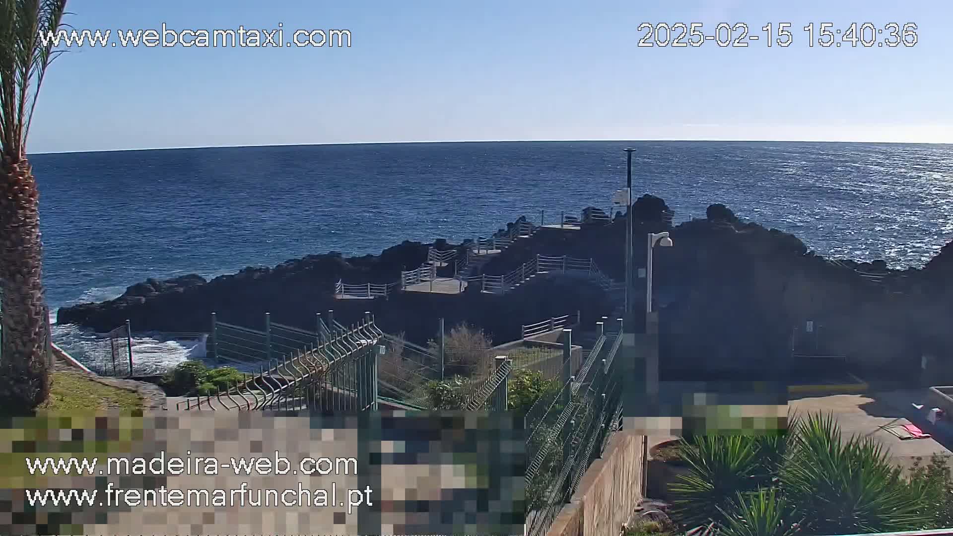 A rocky coastline meets a calm, blue ocean under a clear, sunny sky, with a walkway and railing leading down to the water's edge.