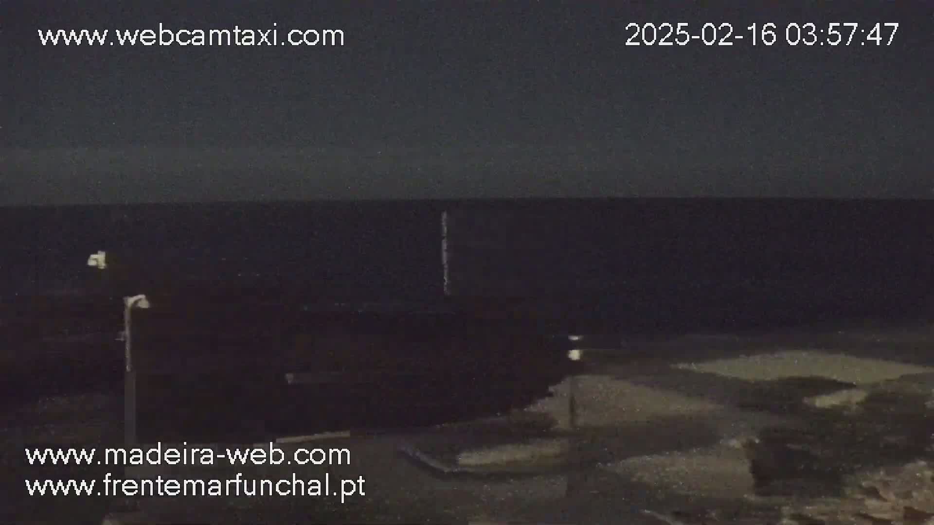A dark, nighttime scene shows a mostly black ocean under a dark sky, with a few lights and poles visible in the foreground.