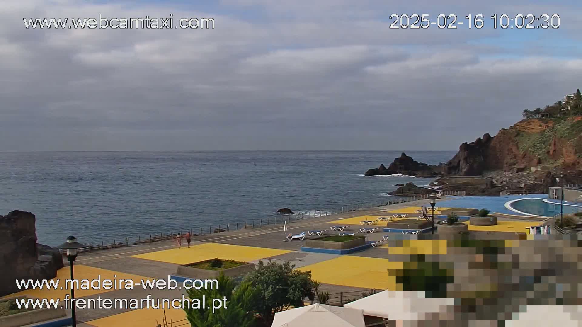 An overcast day reveals a seaside area with a pool, lounge chairs, and yellow-tiled walkways, all set against a backdrop of dark cliffs and a calm ocean.
