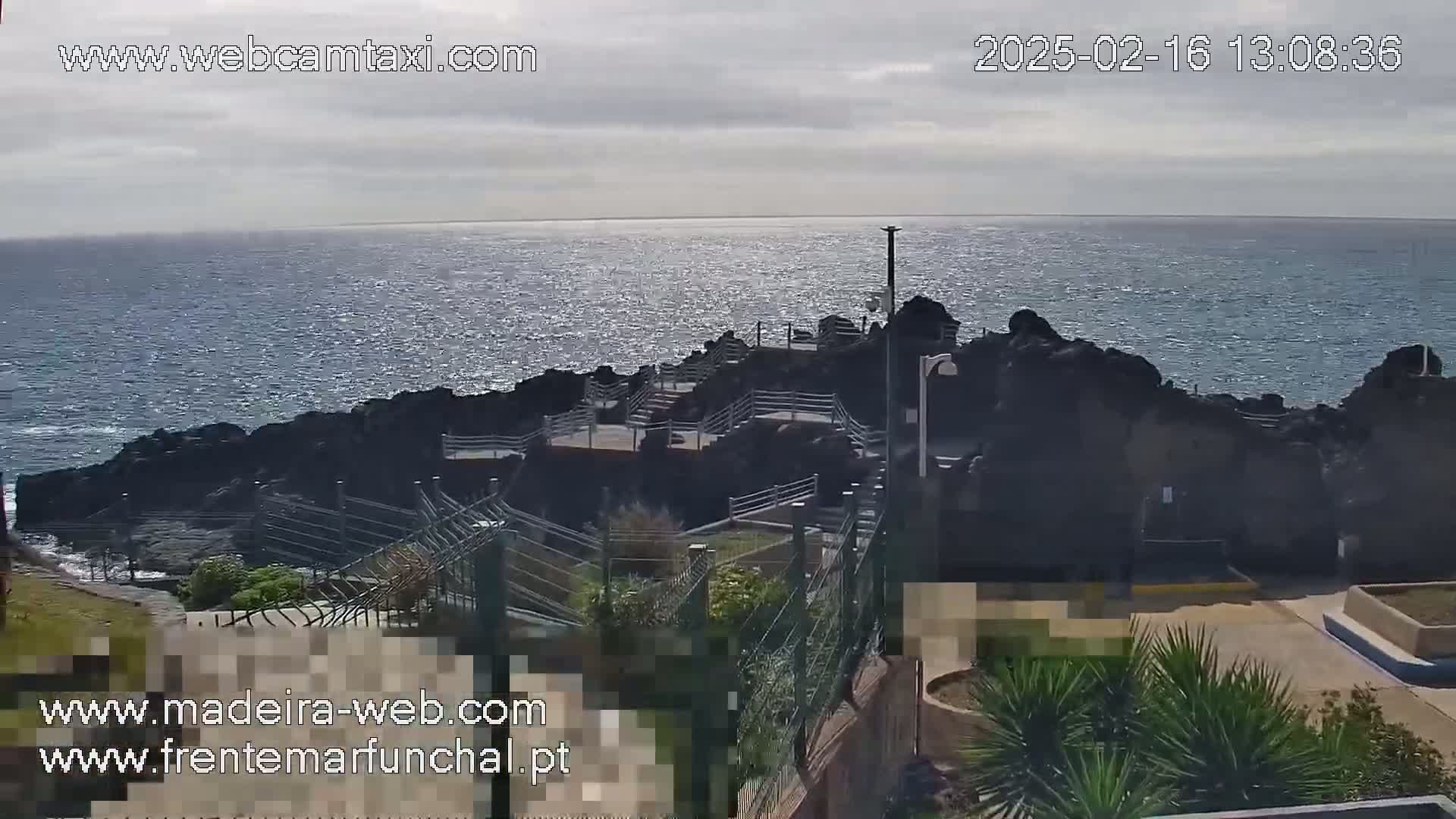 A rocky coastline with a paved walkway overlooks a calm, sparkling ocean under a mostly cloudy sky.