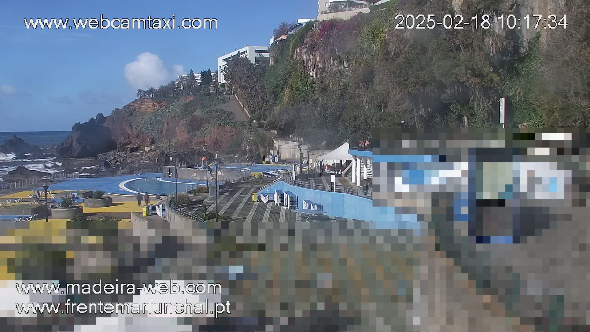 An outdoor scene on a sunny day shows an oceanside pool area with buildings and cliffs in the background.