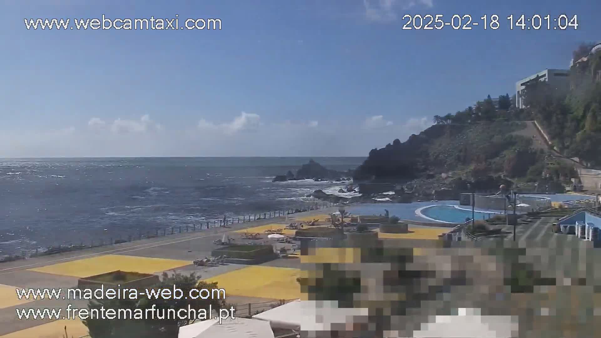 An outdoor view of a seaside pool area with a mostly sunny sky and calm ocean waves.