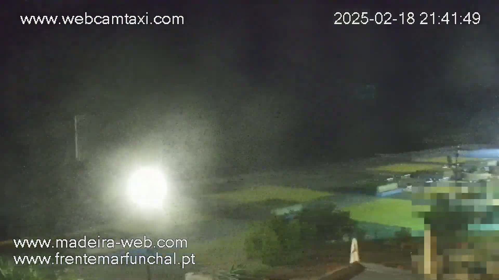 A nighttime, hazy outdoor scene shows a brightly lit area near a body of water, with some vegetation visible.