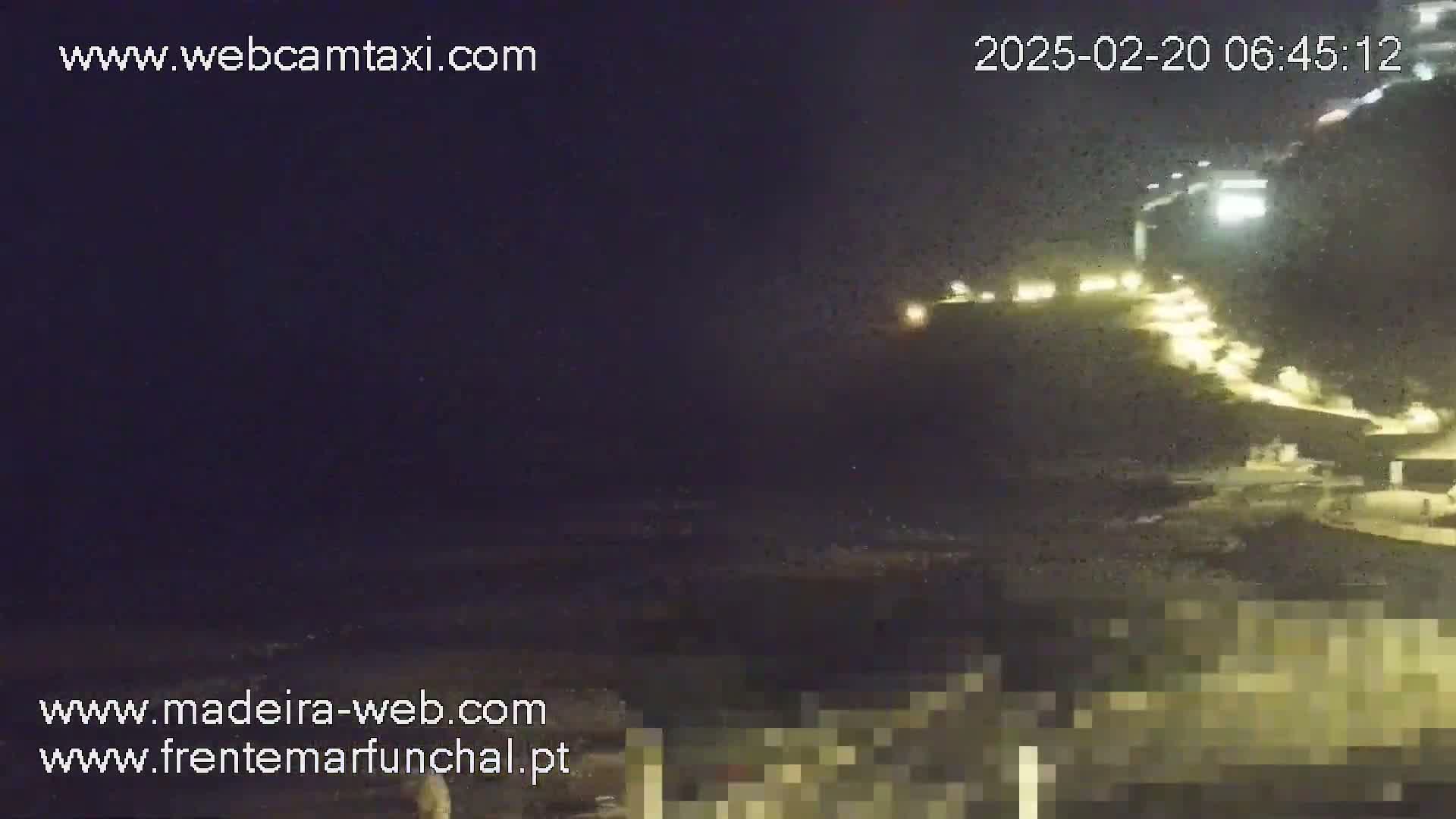 A nighttime coastal scene shows a dark ocean, a brightly lit hillside, and a blurred foreground.