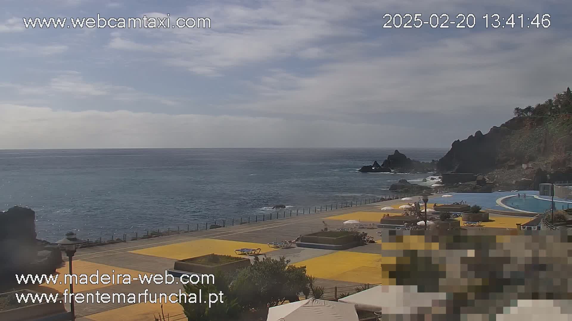 An ocean view on a partly cloudy day shows a paved area with lounge chairs and umbrellas near an outdoor pool adjacent to dark volcanic rock cliffs.
