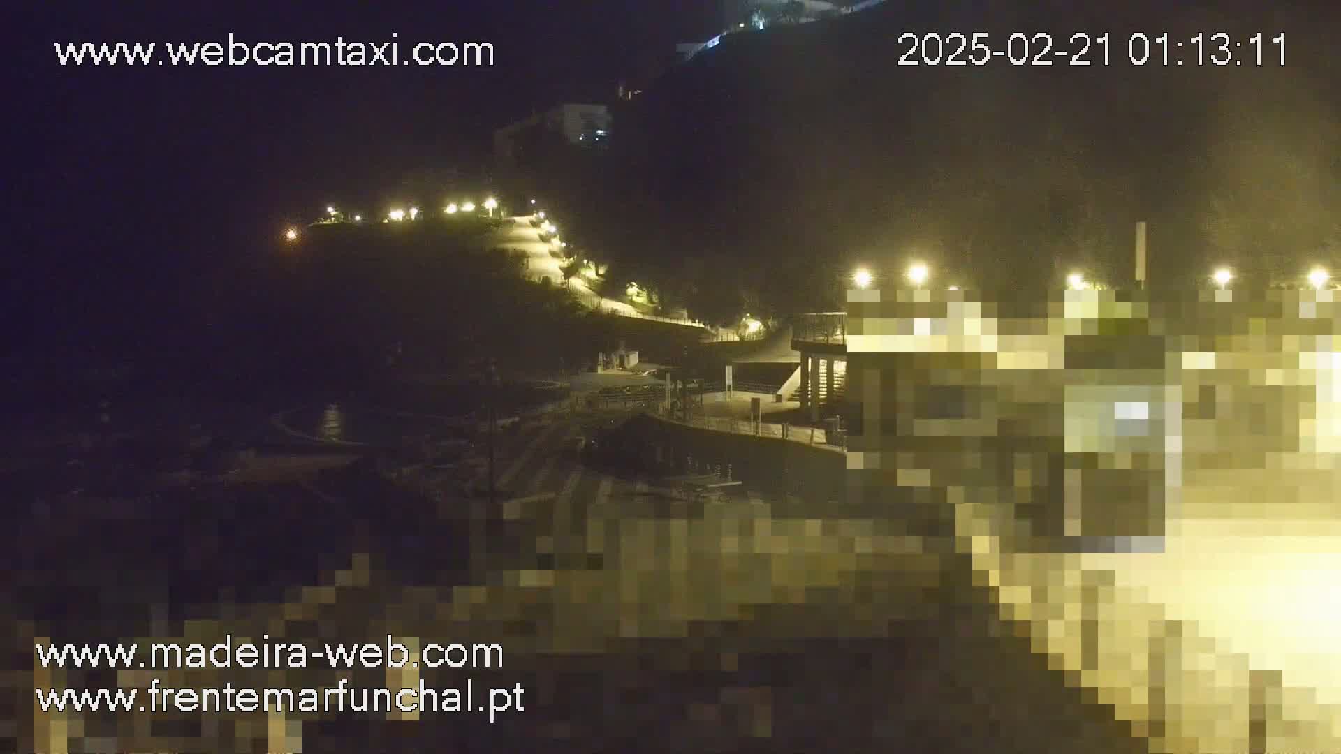 A nighttime, coastal scene shows a paved walkway illuminated by lights winding down a hillside toward a dark ocean with waves, ending at a building complex also lit up.