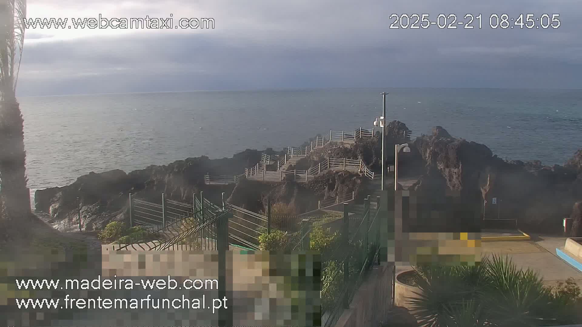 A rocky coastline with a walkway and railing overlooks a calm ocean under an overcast sky.