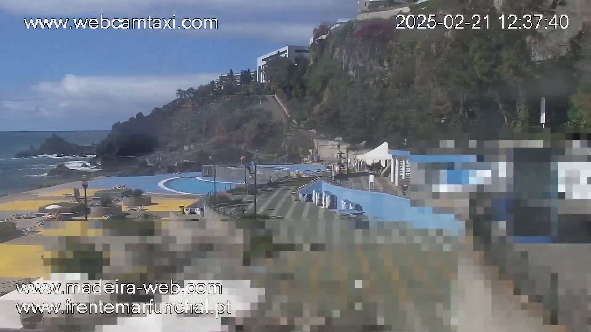 A sunny outdoor scene shows a seaside pool area with a building and a rocky coastline.