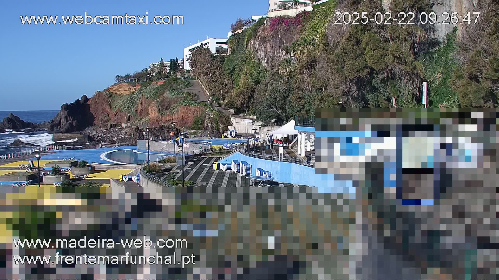 An outdoor scene on a sunny day shows an oceanside swimming pool complex situated on a cliff overlooking the ocean.
