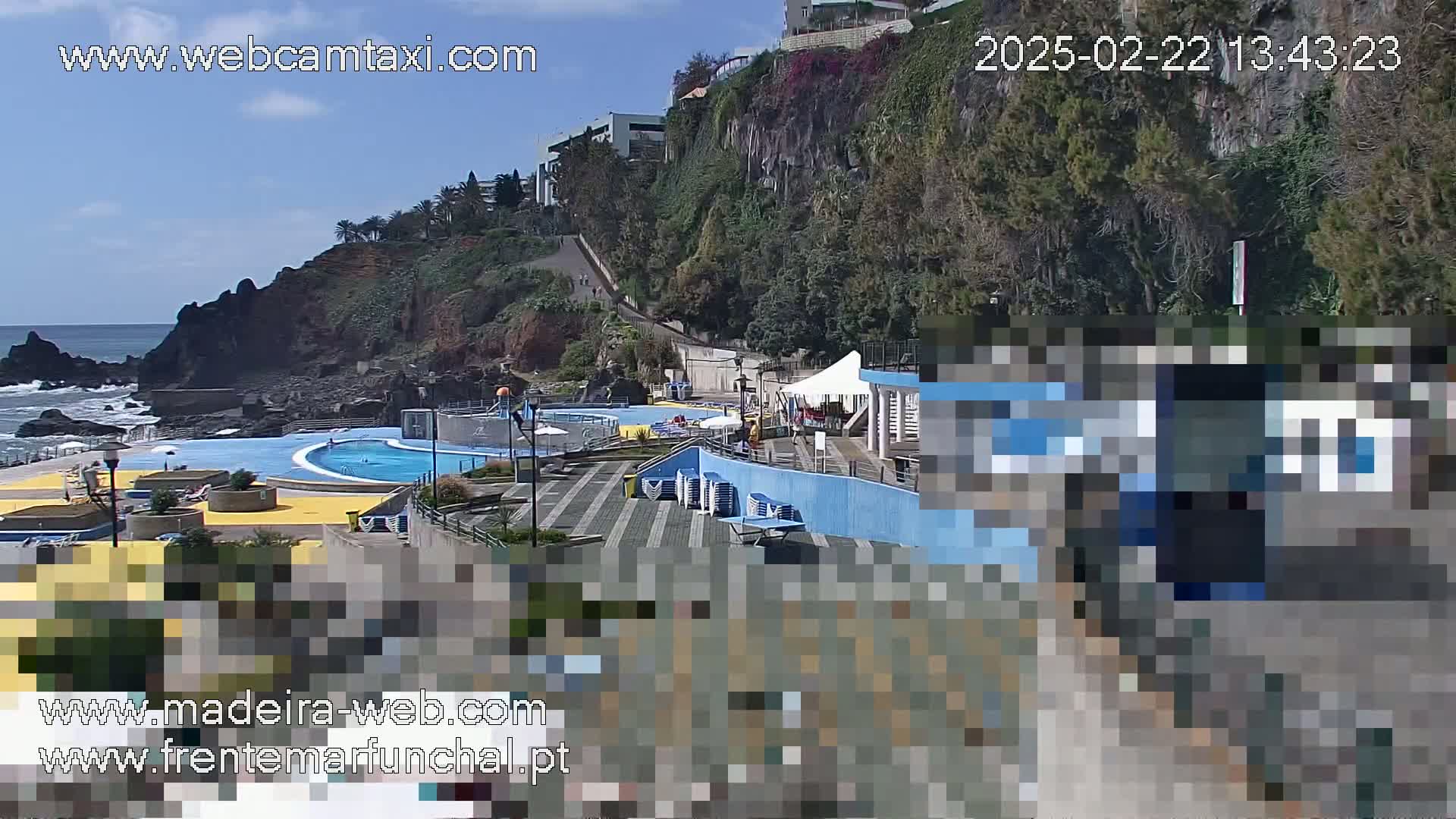 An outdoor scene on a sunny day shows a seaside pool complex situated on a rocky cliff overlooking the ocean.