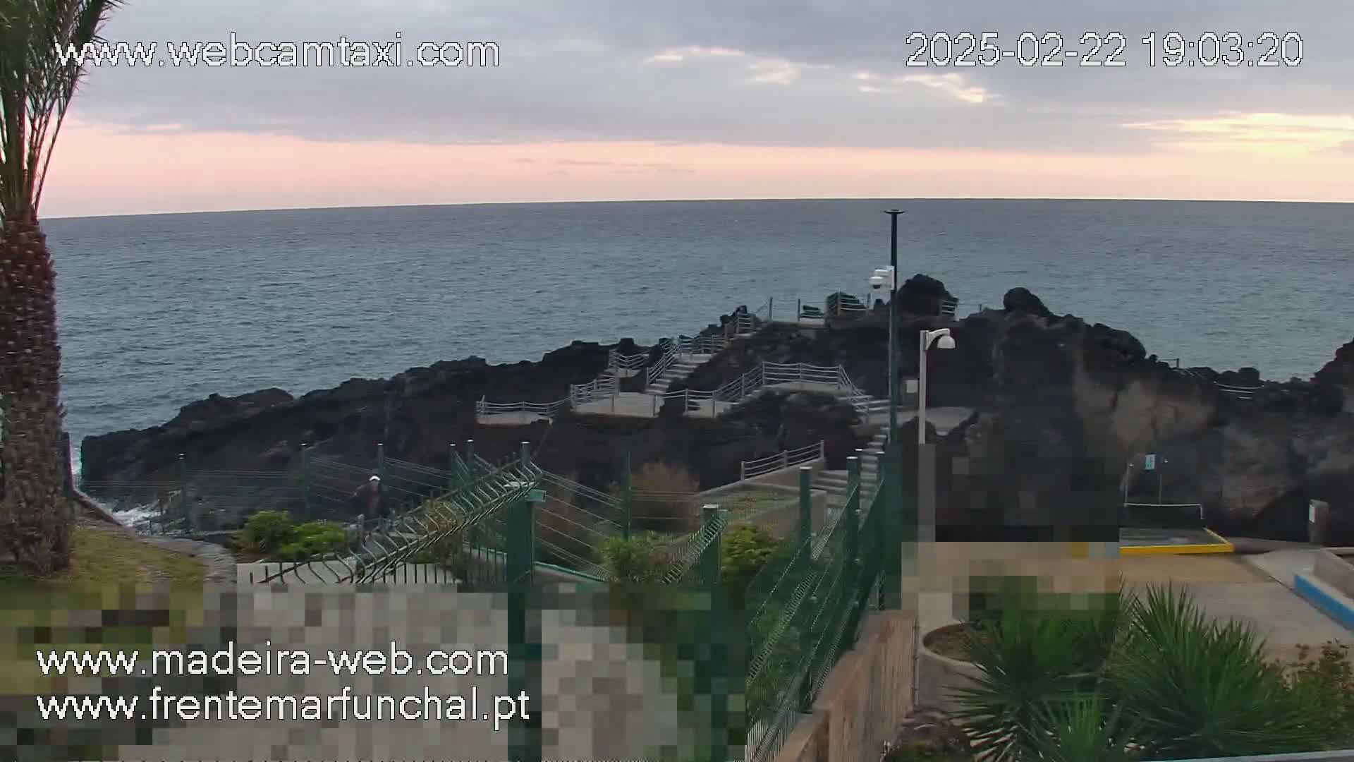 A rocky coastline with stairs leading down to the ocean is visible under an overcast sky at dusk.
