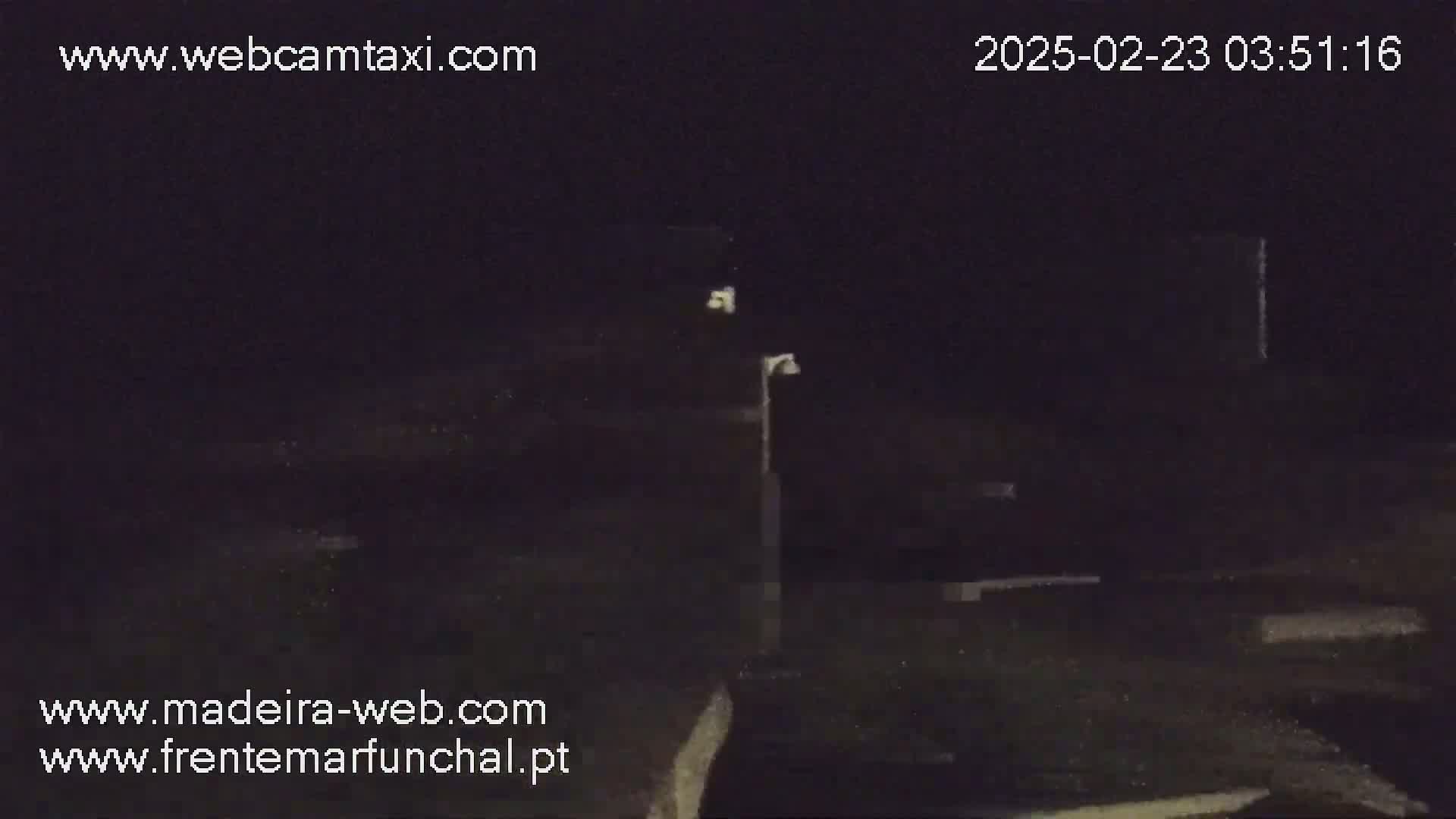 A nighttime, low-light image shows a dark, indistinct outdoor scene with a single light post and some vaguely defined structures.
