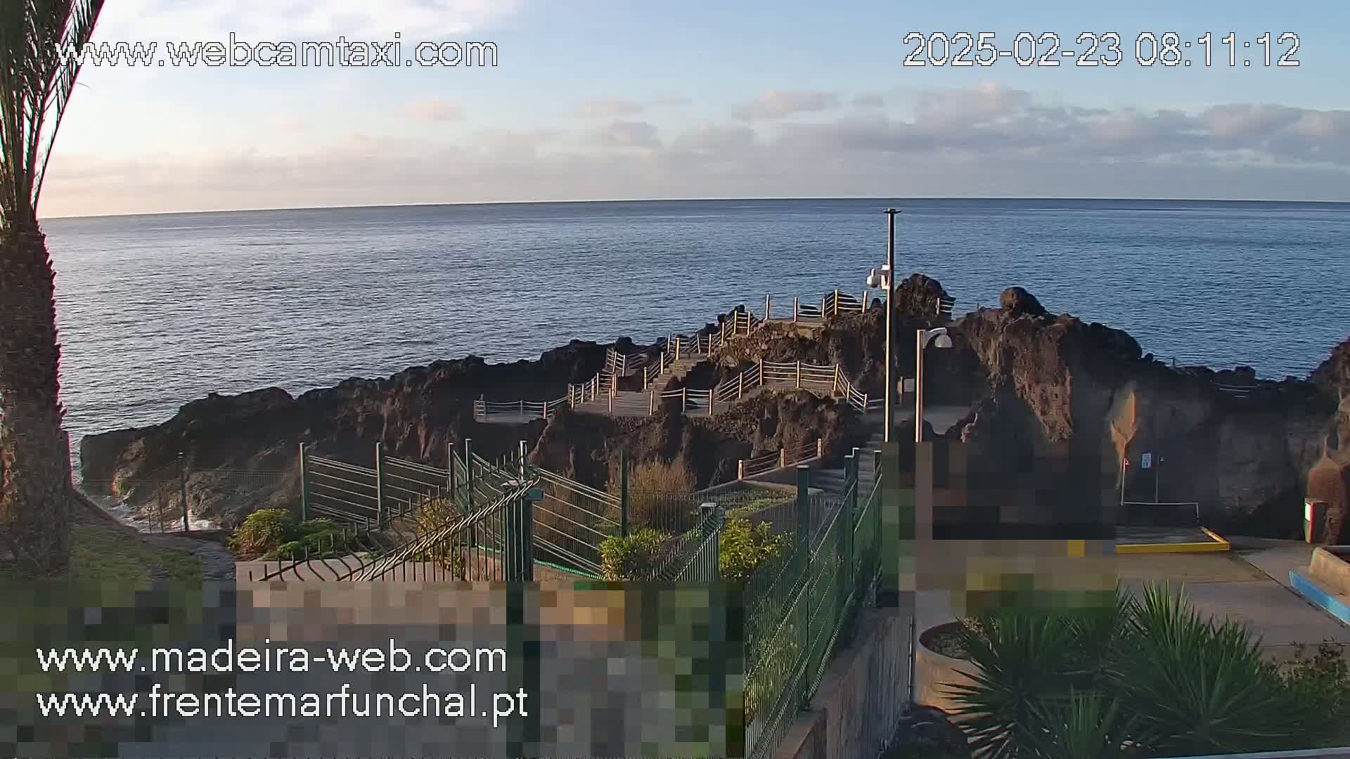 A rocky coastal area with stairs and railings overlooks a calm ocean under a partly cloudy sky.