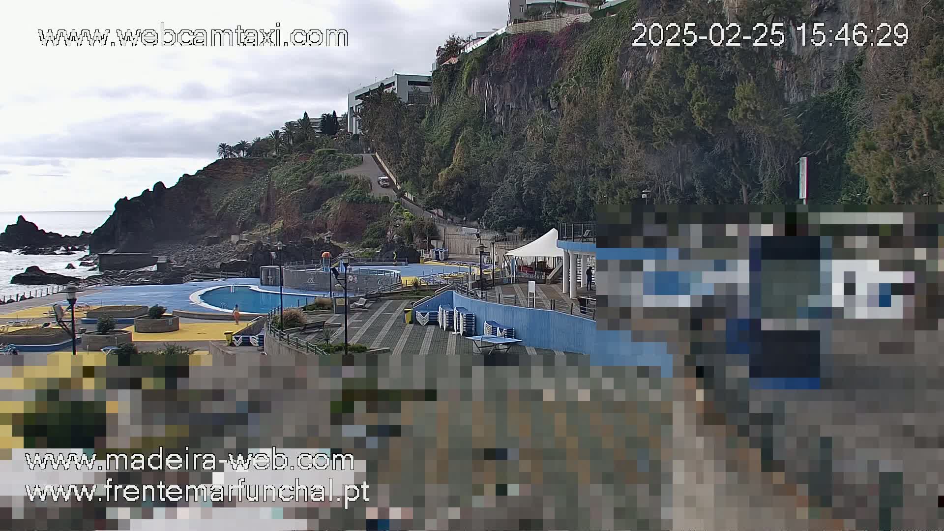 An outdoor scene on an overcast day shows an oceanside pool complex nestled below a steep, tree-covered cliff.