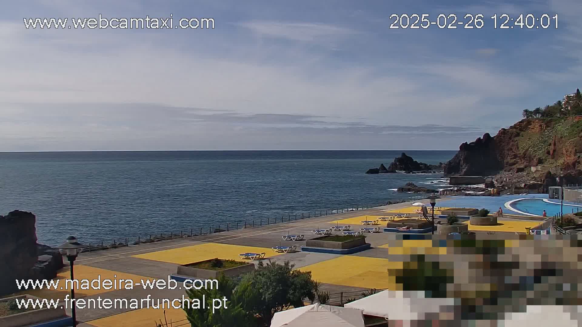 A mostly sunny outdoor scene shows a calm ocean next to a paved area with lounge chairs and a swimming pool, all situated against a rocky cliff.