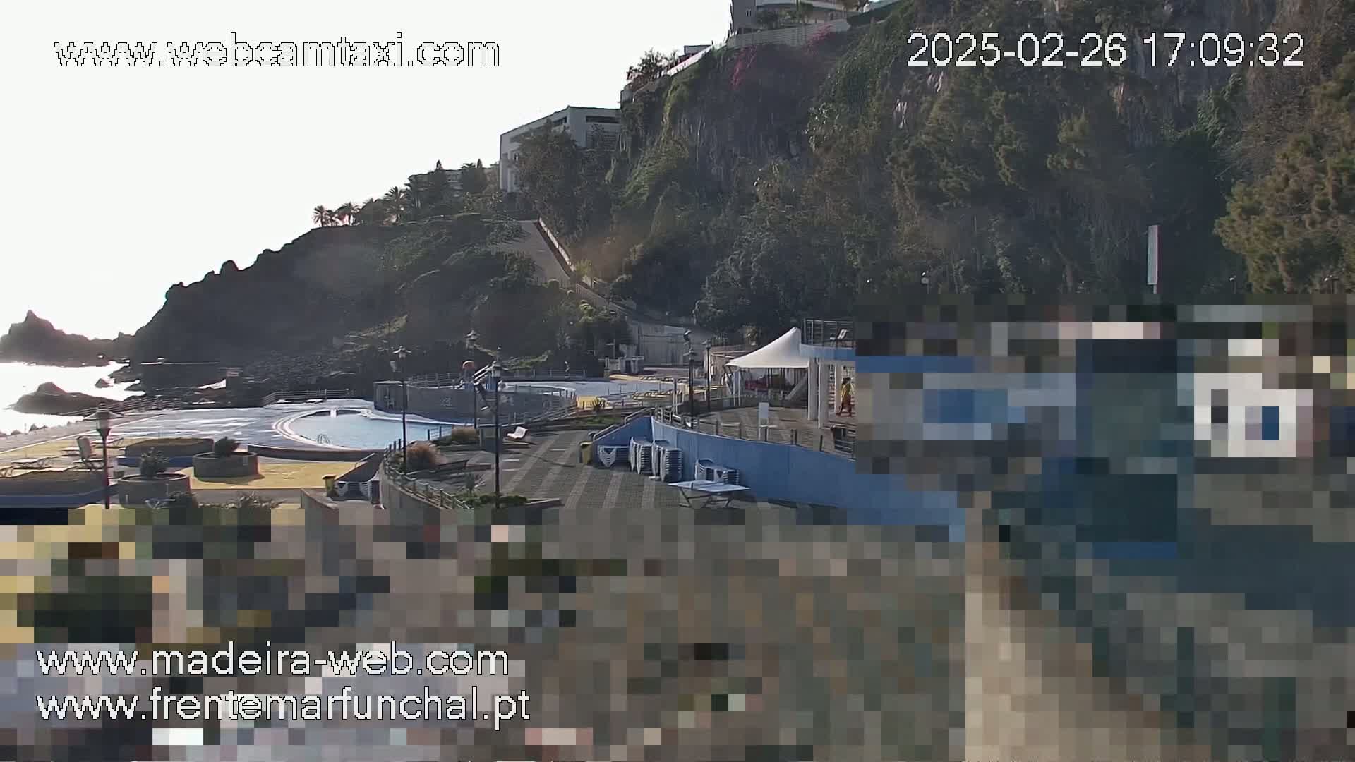 An outdoor view of a seaside pool area next to a rocky cliff, under a mostly clear sky.