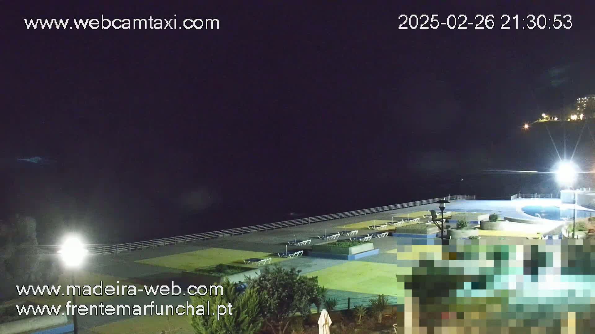 A nighttime view of a dark ocean, a pool area with lounge chairs, and some landscaping, illuminated by artificial lights.