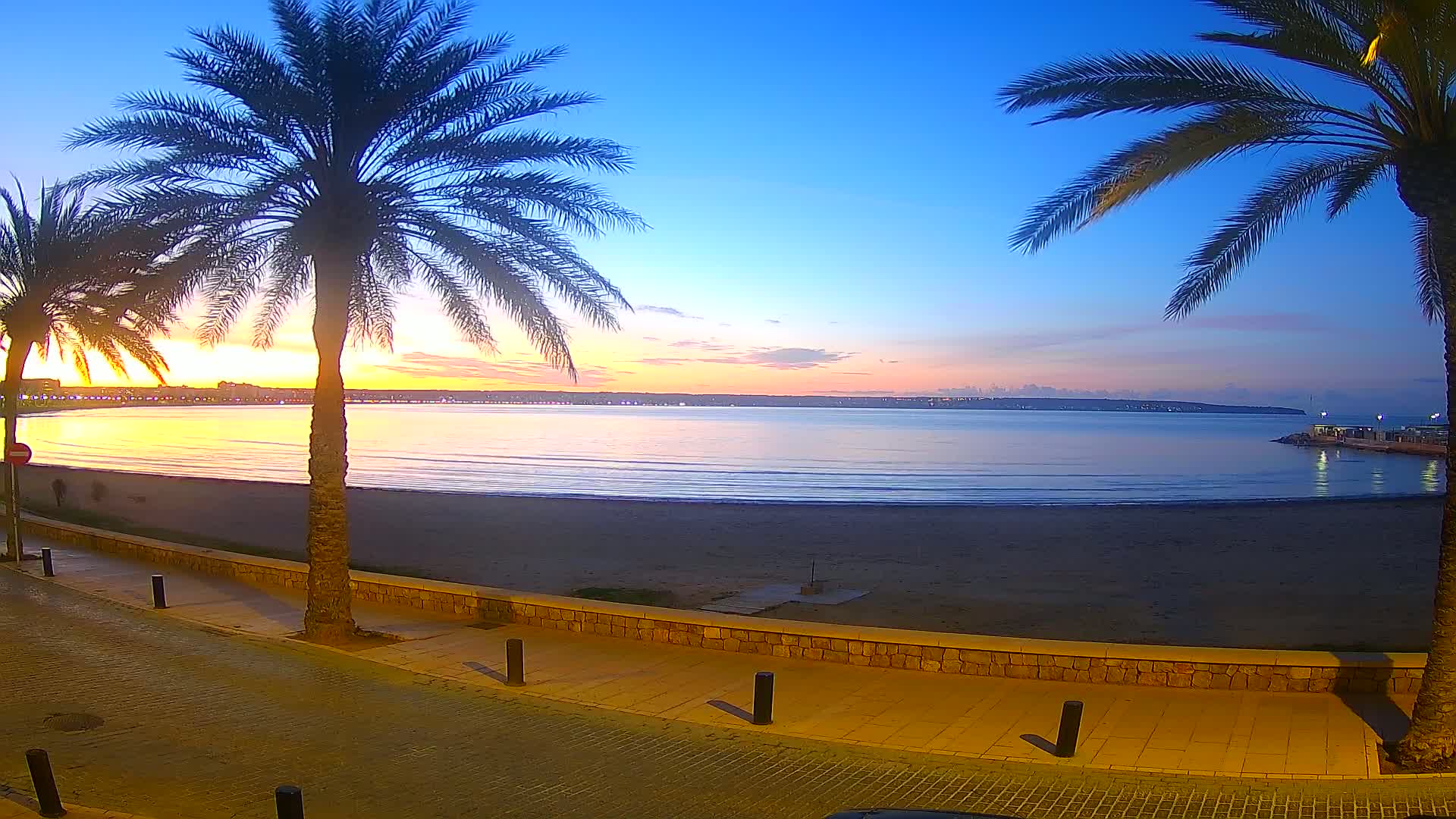 A sandy beach at sunrise, bordered by palm trees and a paved walkway, overlooks a calm sea with a distant city skyline.