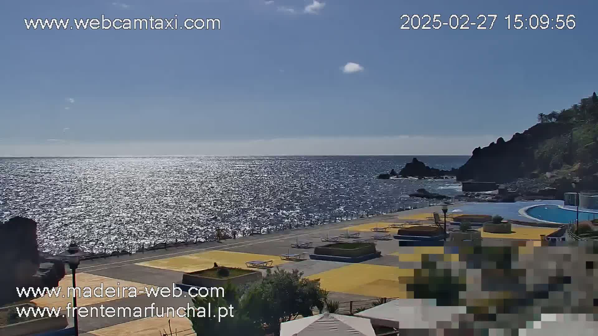 A sunny day overlooking a sparkling ocean with a swimming pool and lounge chairs situated on a paved area next to dark volcanic rock formations.