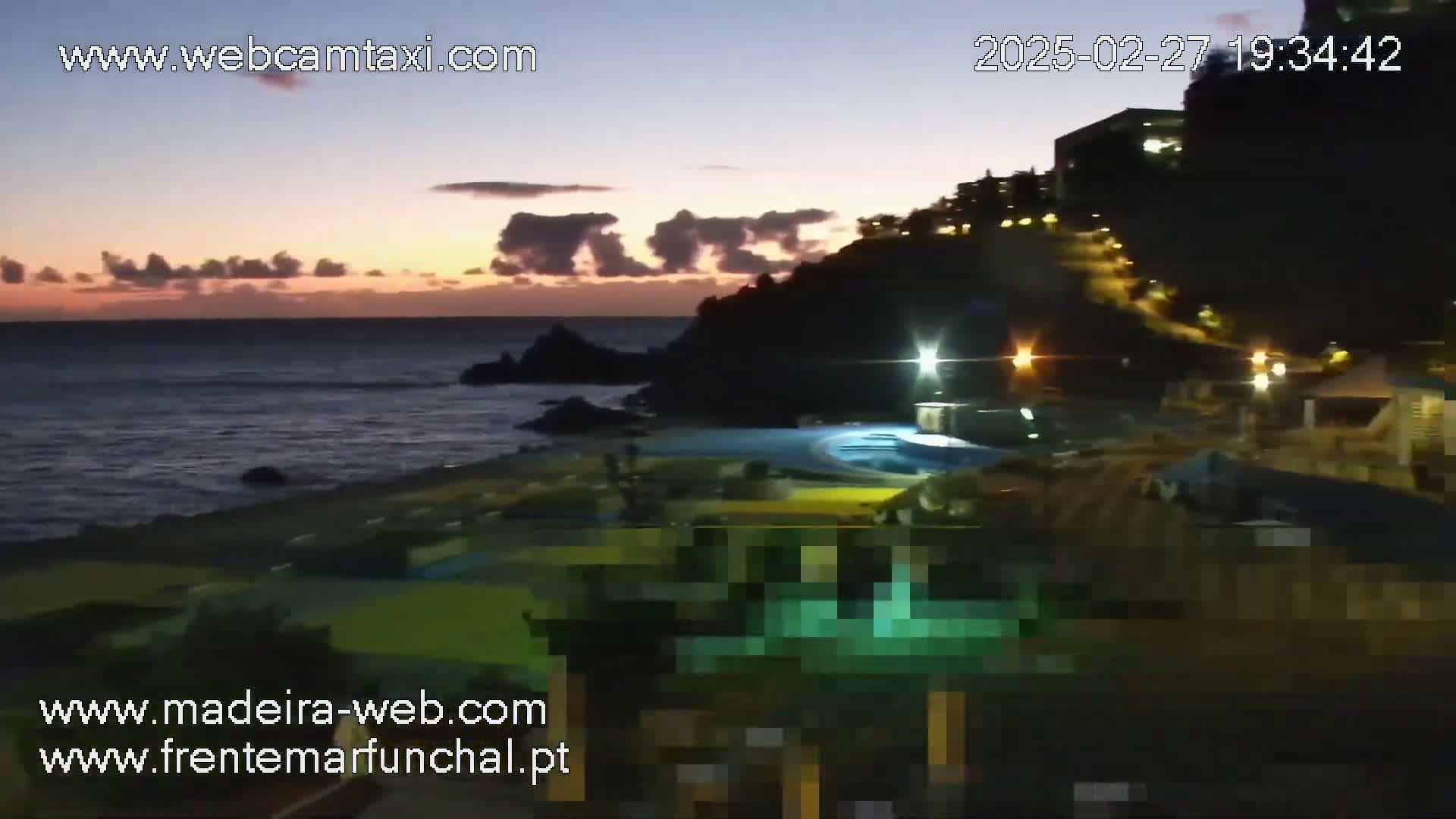 A dark, coastal scene at twilight shows a body of water under a partly cloudy sky, with some buildings and lights visible on a cliffside at night.