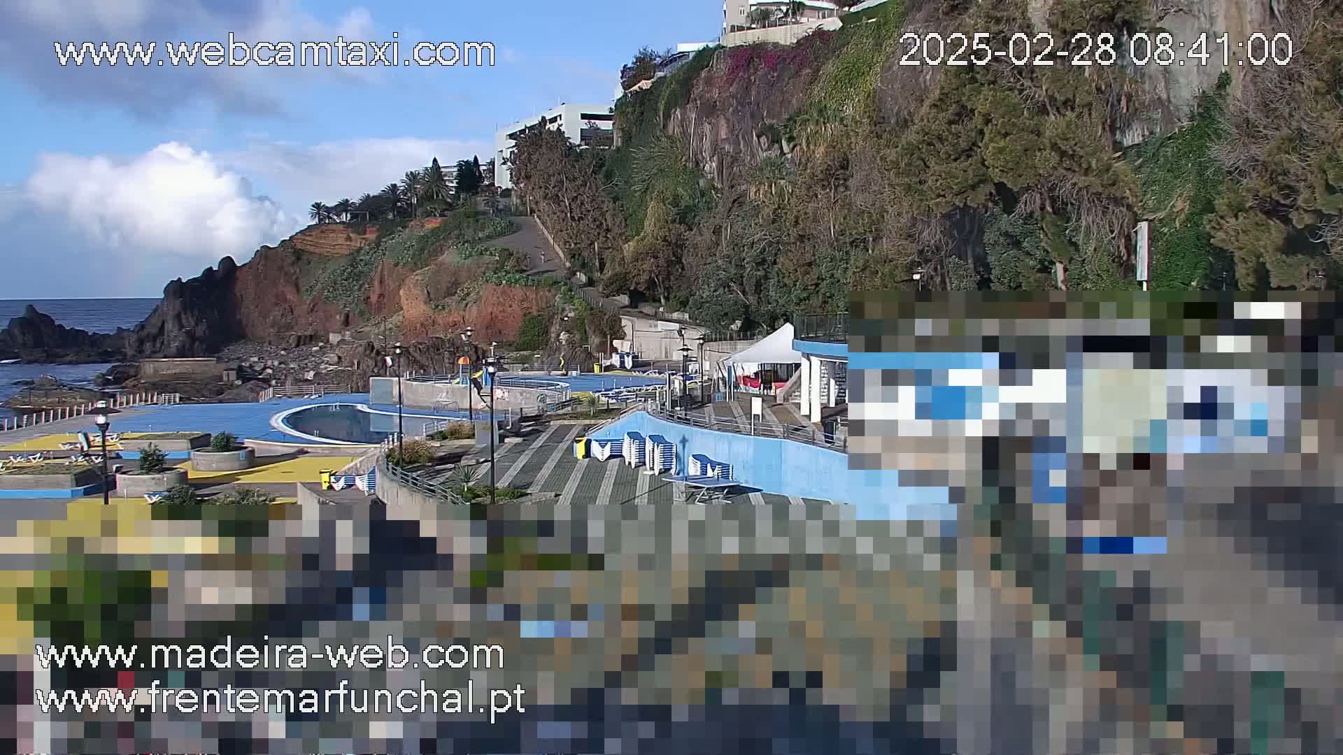 An outdoor view of an oceanside pool complex nestled against a rocky cliffside on a partly cloudy day.