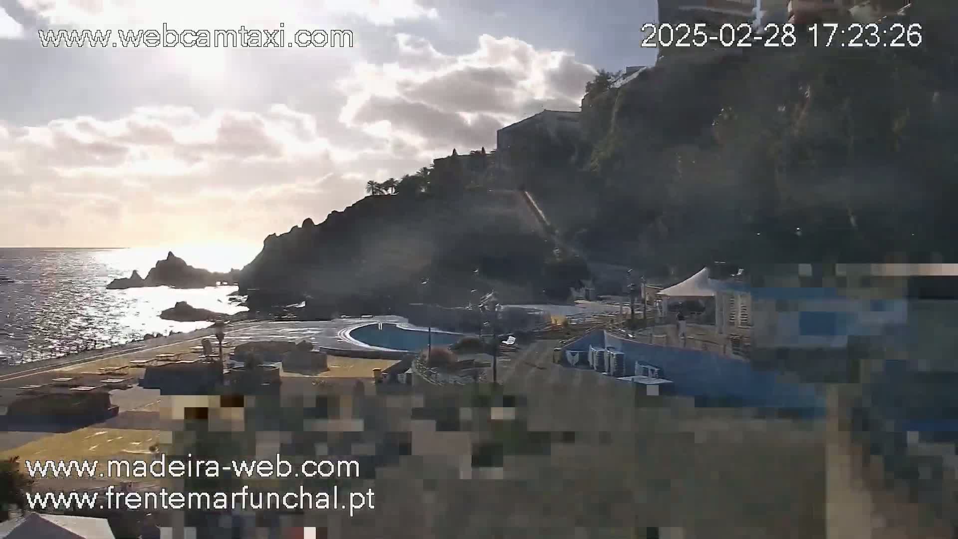 A partly cloudy sunset over a rocky coastline with a swimming pool and buildings visible near the water.