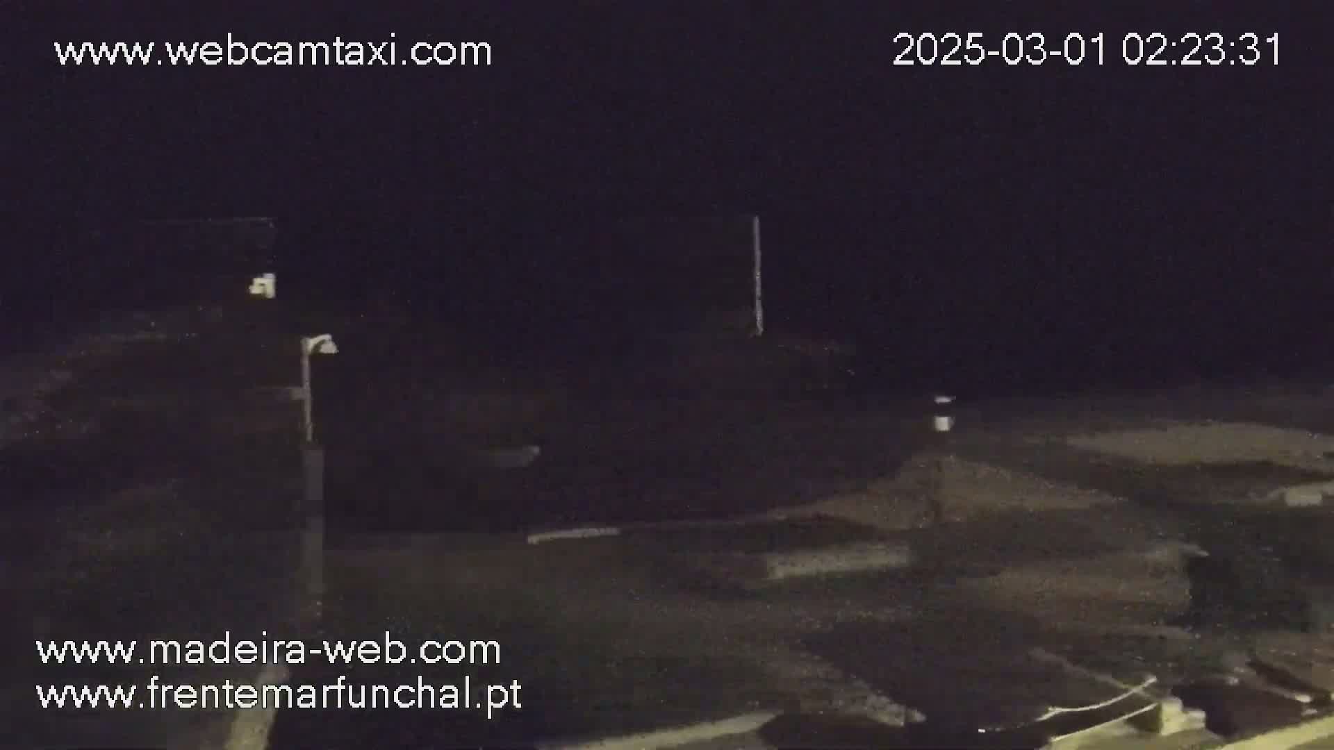 A dark, nighttime outdoor scene shows a mostly empty paved area with a few indistinct objects and a single lamppost.