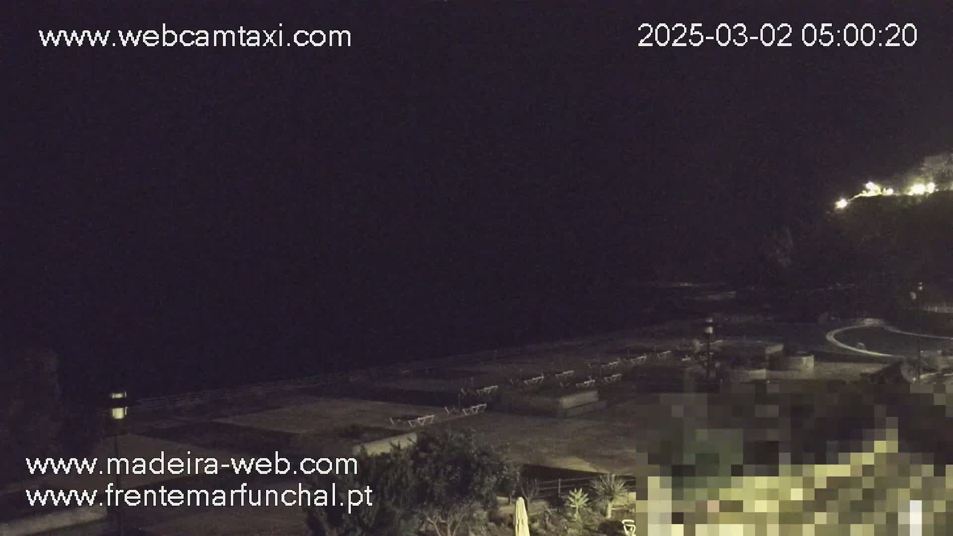 A nighttime, low-light view of an empty outdoor area with lounge chairs near a body of water, under a dark sky.