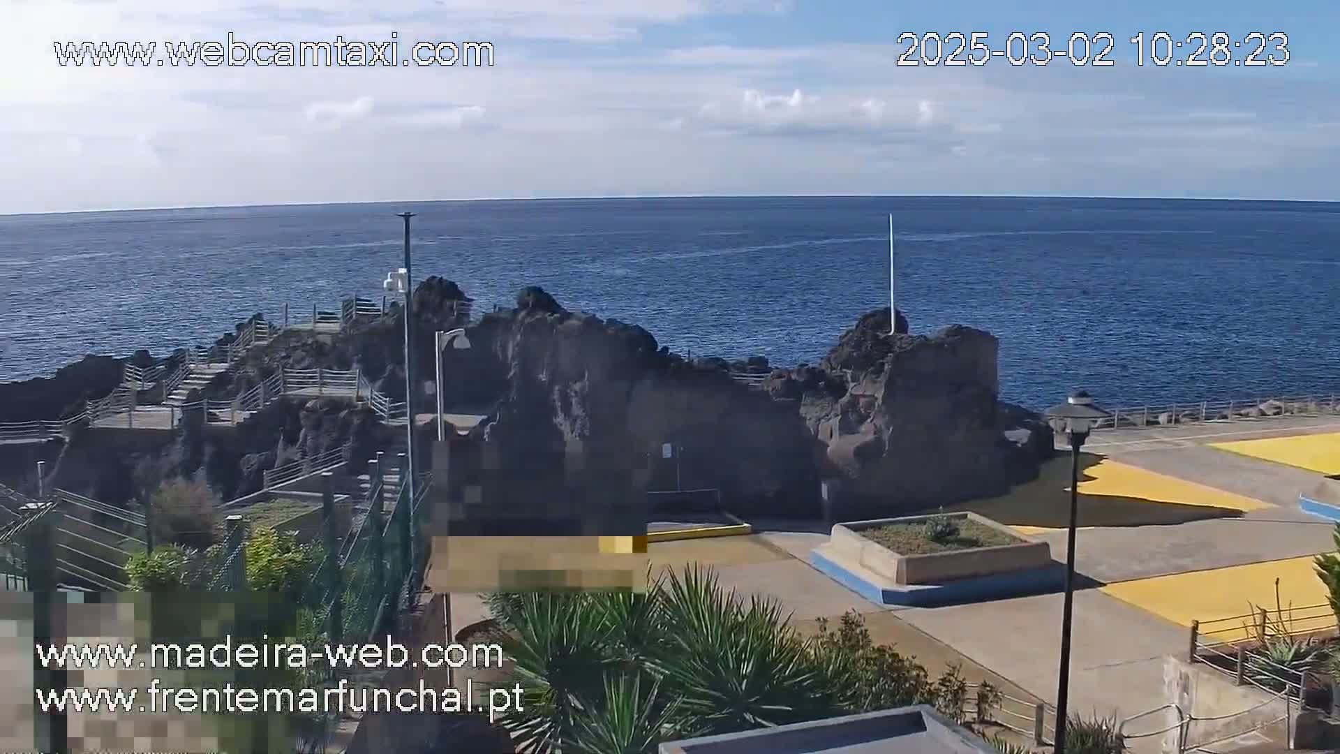 A partly cloudy day reveals a rocky coastline with a paved area featuring yellow sections and low-lying vegetation, overlooking a calm, dark blue ocean.