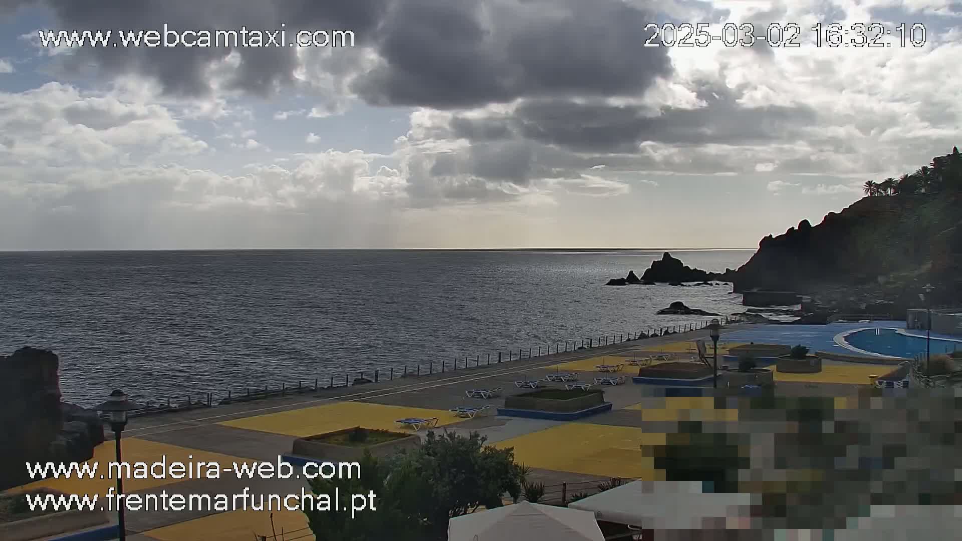 A partly cloudy day overlooks a dark-blue ocean with a coastline featuring a paved area with lounge chairs surrounding a pool.