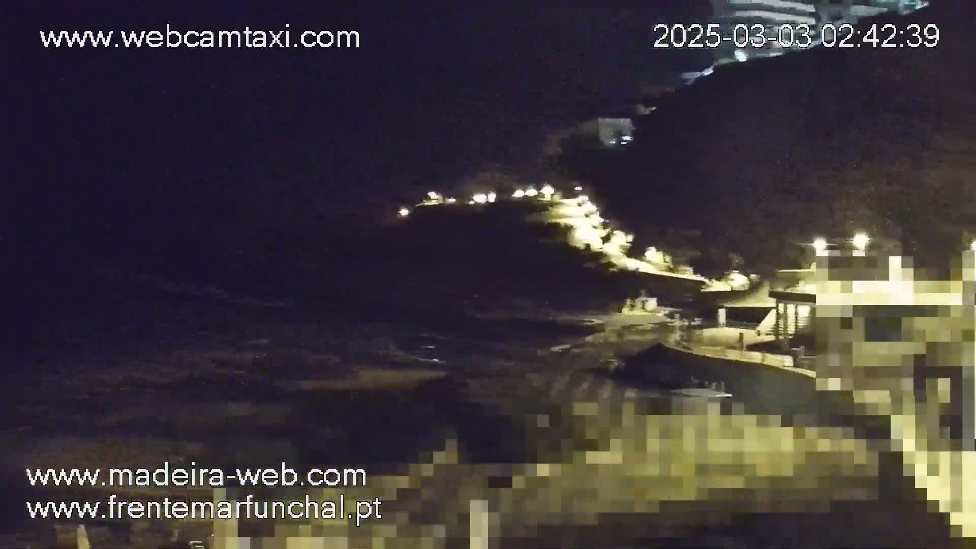 A nighttime view of a coastline with dark, wavy water, illuminated buildings on a hill, and lights along a road.